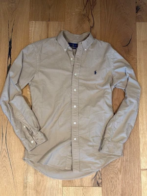Beige skjorta från Polo Ralph Lauren - Klassisk beige skjorta från Polo Ralph Lauren med mörkblå broderad logga på bröstet. Skjortan har knappar framtill och på manschetterna samt en tidlös passform. Perfekt till både jeans och chinos.