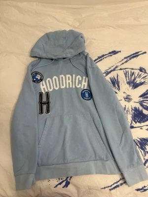 Ljusblå hoodie från Hoodrich - Säljer en ljusblå hoodie från Hoodrich med vita och svarta broderade detaljer och patches på bröstet. Hoodien har stor magficka, dragsko i huvan och ribbade muddar. Perfekt för dig som gillar streetwear och vill sticka ut med snygga detaljer.