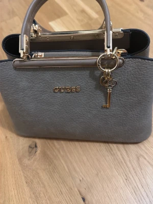 Grå handväska från Guess - Snygg grå handväska från Guess med guldfärgade detaljer och ett dekorativt nyckelberlock. Väskan har dubbla handtag, strukturmönstrat konstläder och dragkedja upptill. Perfekt accessoar för att lyfta din outfit.