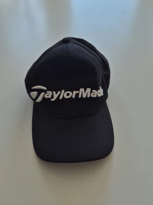 Svart TaylorMade keps - Snygg svart keps från TaylorMade med broderad vit logga framtill. Klassisk böjd skärm och justerbar baksida. Perfekt för dig som gillar sportig stil och vill ha en stilren accessoar till vardagslooken.