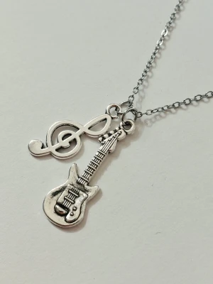 Halsband med gitarr och nota - Snyggt halsband i silverfärgad metall med två hängen: en elgitarr och en g-klav. Perfekt för dig som älskar musik och vill ha en unik accessoar som sticker ut. Kedjan är supertunn men slitstark och smidig, och detaljerna på motiven är tydliga och coola. Materialet rostfritt stål 