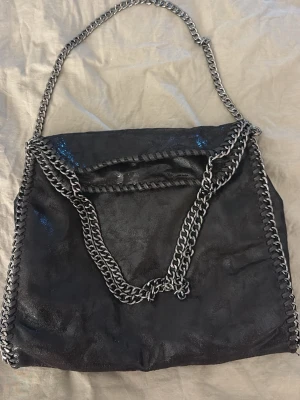 Svart handväska med kedjedetaljer - Snygg svart handväska i mjukt material med silverfärgade kedjor som axelrem och dekor längs kanterna. Väskan har ett rymligt huvudfack och innerficka med dragkedja. Perfekt för dig som gillar en edgy och trendig look.
