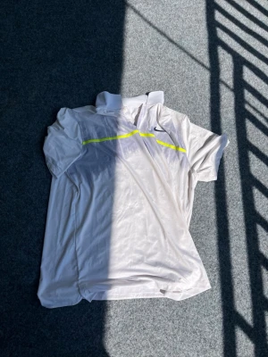 Vit Nike pikétröja med neongul detalj - Säljer en vit sportig pikétröja från Nike med korta ärmar och krage. Tröjan har en neongul linje över bröstet och en liten Nike-logga på vänster sida. Tillverkad i ett lätt och snabbtorkande material, perfekt för träning eller tennis.