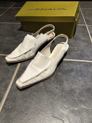 Vita loafers med öppen häl från Oscaria - Säljer ett par vita loafers från Oscaria i mjukt skinn med öppen häl och fyrkantig tå. Skorna har dekorativa sömmar runt tån och små hål på ovandelen för extra stil. Perfekta för dig som gillar en clean och modern look. I storlek 37. Använda en gång.