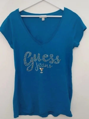 Blå t-shirt Guess Jeans med strass - Snygg blå t-shirt från Guess Jeans med v-ringning och korta ärmar. Framsidan har ett stort Guess Jeans-tryck i strass som ger extra bling. Perfekt till jeans eller kjol för en cool och trendig look. I storlek L