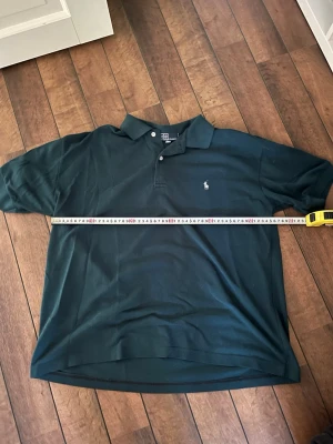 Mörkgrön pikétröja från Polo Ralph Lauren - Klassisk mörkgrön pikétröja från Polo Ralph Lauren med vit broderad logga på bröstet. Tröjan har krage, korta ärmar och knäppning med två knappar framtill. Perfekt för en stilren och avslappnad look. Står ej någon storlek i tröjans nacke därav finns mått.