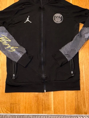 Svart PSG x Jordan ziptröja - Snygg svart ziptröja från PSG x Jordan med vita och grå detaljer. Tröjan har tryckt Paris Saint-Germain-logga på bröstet, Jordan-logga och grafiskt mönster på ärmarna samt texten 'Paris' i gult. Perfekt för dig som gillar sportig streetwear.