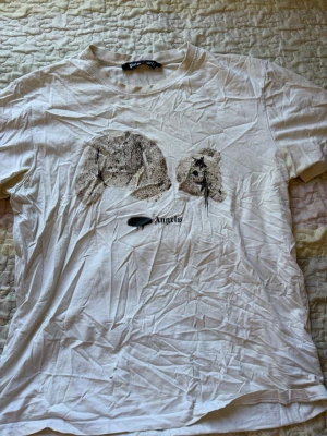 Vit t-shirt med tryck från Palm Angels - Vit t-shirt från Palm Angels med ett stort tryck av ett nallehuvud på bröstet och texten 'Angels' under. Klassisk rund hals och korta ärmar. Tillverkad i mjuk bomull som känns skön mot huden. Perfekt för dig som gillar streetwear och coola prints.