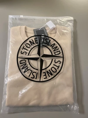 Beige sweatshirt från Stone Island - Stilren beige sweatshirt från Stone Island med rund halsringning och ribbade muddar. Stor Stone Island-logga framtill och klassisk patch på ärmen. Tillverkad i mjukt bomullsmaterial som ger en skön och avslappnad look.