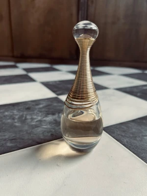 J’Adore Dior 50ml - Säljer en elegant parfymflaska i droppform med transparent glas och guldfärgad spiral runt halsen. Flaskan har en rund, klar glaskula som lock och en stilren, lyxig design som verkligen sticker ut i badrumshyllan. Skick som på bilderna.