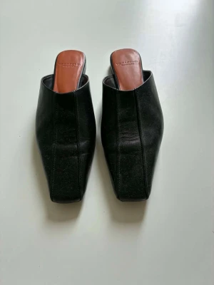 Svarta skinnmules från Vagabond - Stilrena svarta skinnmules från Vagabond i äkta skinn med fyrkantig tå och öppen häl. Snygg minimalistisk design och brunt innersula. Perfekta för dig som gillar en clean och modern look.Använd 1 gång såsom nya. Stor i strl så mer som 37:a . Inköpt sommaren 2025 för 1299:-