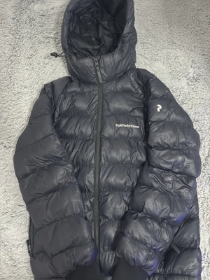 Svart dunjacka Peak Performance - Snygg svart dunjacka från Peak Performance med huva och dragkedja framtill. Jackan har puffig design, diskret logga på bröstet och är tillverkad i glansigt material. Perfekt för kalla vinterdagar och håller dig varm hela säsongen.