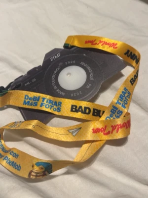 Bad Bunny World Tour kamerahalsband - Unikt halsband från Bad Bunny World Tour med en kameraformad dekoration i svart och grått. Bandet är gult med färgglada tryck och text som 'DeBi Tirar Mas Fotos' och 'Bad Bunny'. Perfekt accessoar för fans och samlare.
