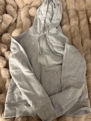 Ljusgrå zip-hoodie med huva - Ljusgrå hoodie med dragkedja och huva, tillverkad i mjukt och bekvämt material. Modellen har kängurufickor framtill och ribbade muddar vid ärmslut och nederkant. Perfekt för en avslappnad och stilren look.