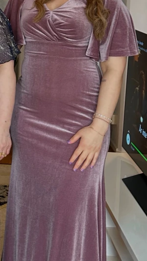Lång lila sammetsklänning - Elegant långklänning i mjuk lila sammet med v-ringning och vida korta ärmar. Klänningen har en markerad midja och ett lätt utställt fall som ger en snygg siluett. Perfekt för speciella tillfällen när du vill känna dig extra fin.