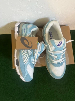 Asics blåvita sneakers - Snygga sneakers från Asics i ljusblått och vitt med mesh och mockadetaljer. Skorna har en sportig look med tydliga Asics-loggor på sidan och tungan. Yttersulan är vit och grå med bra grepp. Perfekta för dig som gillar stilrena och bekväma sneakers.