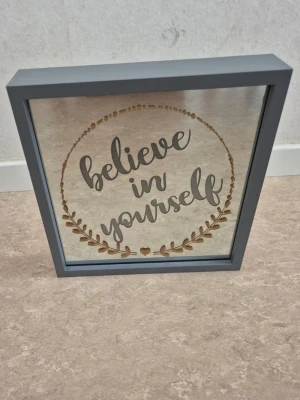 Grå dekorativ tavla med text - Snygg fyrkantig tavla med grå ram och spegelliknande front. Tavlan har texten 'believe in yourself' i svart och guld med dekorativ krans runt. Perfekt som peppande inredningsdetalj till rummet eller kontoret. Går på batteri och har varmvit belysning som lyser upp texten.