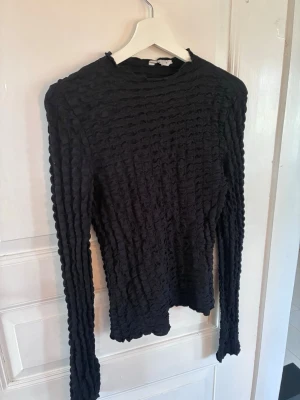 Svart långärmad topp med struktur Cubus - Säljer en svart långärmad topp med coolt strukturerat tyg och rund hals. Toppen har en tight passform och är perfekt för dig som gillar en unik look. Passar till många olika stilar och är enkel att matcha med andra plagg.