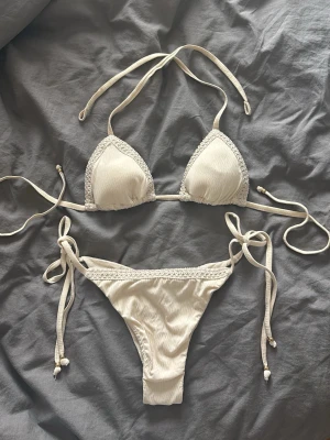 Vit triangelbikini från Calzedonia  - Snygg vit bikini i triangelmodell med knyt både i ryggen, nacken och på sidorna av bikinitrosan. Både överdel och underdel har fina virkade detaljer längs kanterna. Bikinin är från Calzedonia. Kräm vit bikini. Båda delarna följer med såklart. Bikinin kostar 838. 