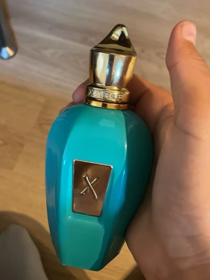 Xerjoff turkos parfymflaska - Unik parfymflaska från Xerjoff i en snygg turkos färg med gulddetaljer och en elegant, geometrisk form. Flaskan har en guldfärgad kork med märkets logga ingraverad och en metallplatta med ett X på framsidan. Perfekt för dig som gillar exklusiva dofter och lyxig design.