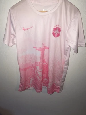 Ljusrosa Nike fotbollströja - Säljer en ljusrosa fotbollströja från Nike med korta ärmar. Tröjan har ett tryck av ett svärd och ett klubbmärke med stjärnor på bröstet. Materialet är lätt och andas, perfekt för träning eller match. Snygg och unik design med rosa detaljer.