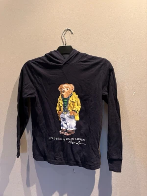 Svart hoodie med Polo Bear-tryck - Svart hoodie från Ralph Lauren med det ikoniska Polo Bear-trycket på bröstet. Björnen har på sig en gul jacka, randig tröja och jeans. Hoodien har långa ärmar, huva och ribbade muddar. Perfekt för dig som gillar streetstyle och vill sticka ut.