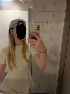 Beige figursydd topp med kort ärm - Snygg beige topp med korta ärmar och figurnära passform. Toppen har en lätt utsvängd nederdel som ger en feminin siluett. Perfekt att styla med jeans eller kjol för en trendig look.
