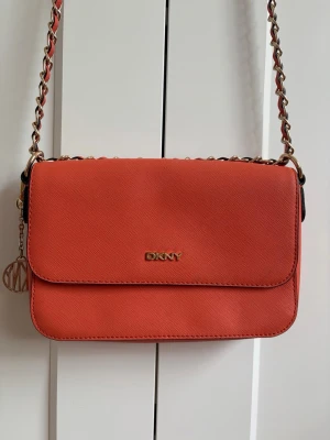 Orange axelväska från DKNY - Snygg orange axelväska från DKNY med guldfärgad kedjerem och logga framtill. Väskan har en stilren, rektangulär form och är tillverkad i ett strukturerat material som ger en exklusiv känsla. Väskans kedja har en liten skada, som man ser i sista bilden. 