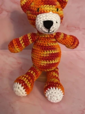 Virkad randig nalle i orange och rött - Söt handvirkad nalle i orange, rött, gult och vitt garn. Nallen har randiga armar och ben, rund mage och ett broderat ansikte med svart nos. Perfekt som dekoration eller present. Sittande 11 cm