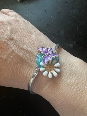 Armband med blomma och fjäril - Unikt armband i silverfärgad metall med en stor vit blomma och en lila fjäril på ett turkost runt fäste. Blomman har gulddetaljer i mitten och armbandet har en öppen design som gör det lätt att ta på och av. Perfekt för dig som gillar färgglada och lekfulla accessoarer.