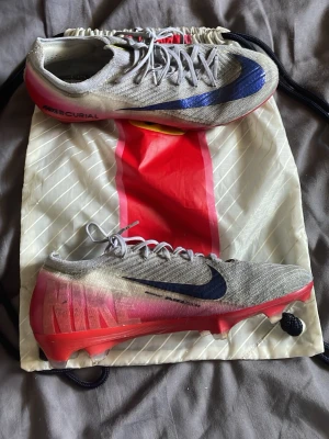 Nike Mercurial fotbollsskor grå/röd - Säljer ett par Nike Mercurial fotbollsskor i grått med blå och svart swoosh samt röda detaljer på sulan. Skorna har låg profil, snörning och är tillverkade i ett lätt syntetmaterial för bästa känsla på planen. Medföljer originalpåse. Tappade bort sulorna