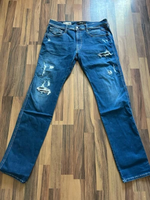 Blå slitna jeans från Replay - Säljer ett par blå jeans från Replay med slitningar och hål för en trendig look. Modellen har raka ben, klassisk femficksdesign och tydliga kontrastsömmar. Perfekta för dig som gillar en avslappnad och cool stil.