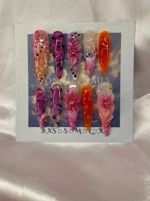 SafariLuxe (XS);ExtraLongStiletto - Set med extra långa handgjorda unika press-on naglar i rosa, lila och orange toner. Naglarna har handmålade blommor, swirl-mönster och små gulddetaljer. Designen är riktigt unik och färgstark, perfekt för dig som vill sticka ut.XS: 14 mm, 11 mm, 11,5 mm, 10 mm, 8 mm