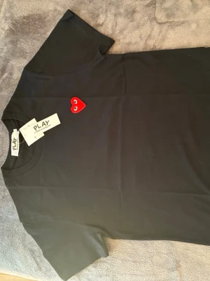 Svart t-shirt med rött hjärta - Snygg svart t-shirt från Comme des Garçons Play med det ikoniska röda hjärtat med ögon broderat på bröstet. Klassisk passform och rund halsringning. Tillverkad i mjuk bomull som känns skön mot huden. Perfekt för dig som gillar stilrena plagg med en twist.