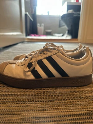 Vita Adidas sneakers med svarta ränder - Säljer ett par klassiska Adidas sneakers i vitt med svarta ränder på sidorna och beige mockadetaljer vid tån. Skorna har lågt skaft, bruna sulor och snörning. Perfekta för dig som gillar retrostil och enkel design.
