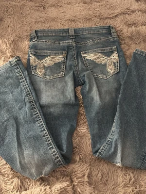 Blå bootcut jeans med broderade fickor nya!! - Säljer ett par blå bootcut jeans med snygga broderade detaljer på bakfickorna. Jeansen har klassisk femficksmodell, normalhög midja och slitningar för en cool vintagekänsla. Materialet är denim i bomull. Perfekta för dig som gillar unik stil!