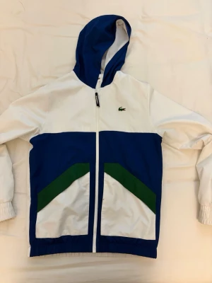 Vit och blå vindjacka från Lacoste - Snygg vindjacka från Lacoste i vitt och blått med gröna detaljer på fickorna. Jackan har huva, dragkedja framtill och elastiska muddar vid ärmslut och nederkant. Klassisk krokodil-logga på bröstet och två stora fickor framtill.