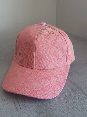 Rosa keps från Gucci med GG-mönster - Snygg rosa keps från Gucci med klassiskt GG-monogram i hela tyget och en liten metallogo framtill. Kepsen har böjd skärm och är tillverkad i ett slitstarkt tygmaterial. Perfekt accessoar för att lyfta din outfit.