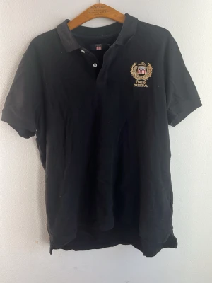Svart pikétröja med broderad Vinson logga - Klassisk svart pikétröja med korta ärmar och krage. På bröstet finns en broderad logga i guld och vitt med texten 'Vision Original'. Tröjan har knappar vid halsen och är tillverkad i ett mjukt material, troligen bomull. Är lite dammig lite överallt därför priset.