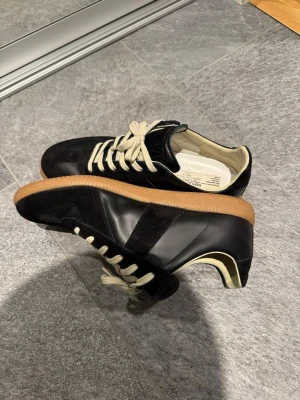 Maison Margiela GATS - Snygga svarta sneakers med beige gummisula och vita snören. Ovandelen är i en mix av läder och mocka för en stilren look. Insidan är ljus och skön, perfekt för dig som gillar klassiska men trendiga skor.