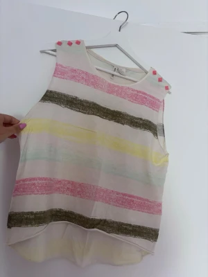 Randig ärmlös linnetopp med pastell - Säljer en söt ärmlös linnetopp med breda pastellfärgade ränder i rosa, gul, grön och vit. Toppen har dekorativa rosa nitar på axlarna och en avslappnad passform med något längre bakstycke. Perfekt för varma dagar och enkel att styla.