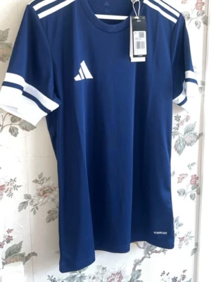 Blå sporttröja från Adidas med lappen kvar  - Snygg blå sporttröja från Adidas med vita detaljer på axlar och ärmar. Tillverkad i ett lätt och snabbtorkande material, perfekt för träning eller match. Klassisk rund halsringning och korta ärmar med vita ränder.