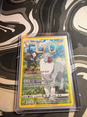 Pokémonkort Wyrdeer - Säljer ett Pokémonkort med Wyrdeer i färgstark design. Kortet har gul ram och motiv av Wyrdeer med blå och gröna naturfärger i bakgrunden. Kortet är inplastat i ett skyddsfodral och har tydliga illustrationer och text.