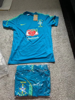 Blå Brasilien fotbollströja Nike - Säljer en blå Brasilien fotbollströja från Nike med CBF-emblem, gula detaljer och Guaraná Antarctica-logga på bröstet. Tröjan har rund hals och korta ärmar, perfekt för träning eller match. Matchande shorts medföljer.