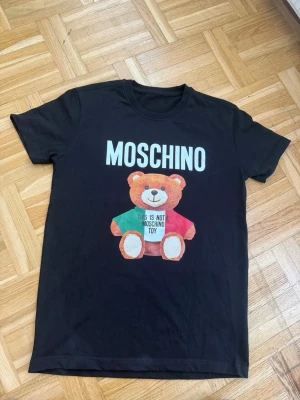 Svart Moschino t-shirt med björntryck - Säljer en svart t-shirt från Moschino med stort tryck av en söt björn och texten 'This is not a Moschino Toy' på framsidan. T-shirten har klassisk passform och korta ärmar. Perfekt för dig som gillar statement-plagg och coola prints. Storlek S helt ny har aldrig använts.