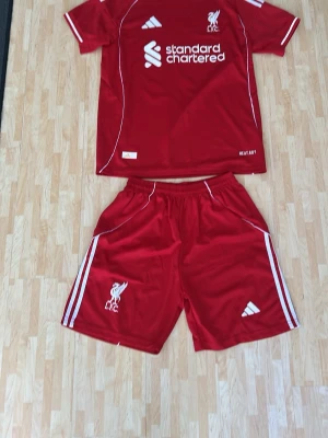 Liverpool röd matchställ Adidas - Säljer ett rött Liverpool matchställ från Adidas med klubbmärke och sponsortryck på tröjan samt shorts. Setet har vita detaljer och klassiska Adidas-ränder på sidorna. Tillverkat i lätt och ventilerande material, perfekt för fotbollsträning eller match.