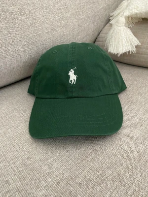 Grön keps från Polo Ralph Lauren unisex - Mörkgrön keps från Polo Ralph Lauren med klassisk vit broderad logga framtill och 'POLO' broderat bak. Justerbart spänne baktill för perfekt passform. Tillverkad i bomullstyg som känns både stilrent och avslappnat.