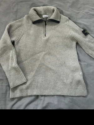 Grå  stickad tröja från Stone Island - Ribbstickad grå tröja från Stone Island med halv dragkedja och hög krage. Tröjan har lång ärm och klassisk Stone Island-patch på ena ärmen. Perfekt för lager på lager och riktigt snygg till jeans. 