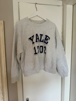 Grå sweatshirt med YALE-tryck - Säljer en ljusgrå sweatshirt med mörkblått YALE tryck på bröstet. Tröjan har rund halsringning, ribbade muddar vid ärmslut och nederkant samt en avslappnad passform. 