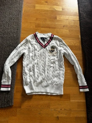 Vit kabelstickad tröja Tommy Hilfiger - Vit kabelstickad tröja från Tommy Hilfiger i premium bomull. Tröjan har V-ringning med röd och marinblå rand, broderad logga på bröstet och ribbade muddar med matchande ränder. Perfekt för en preppy look.
